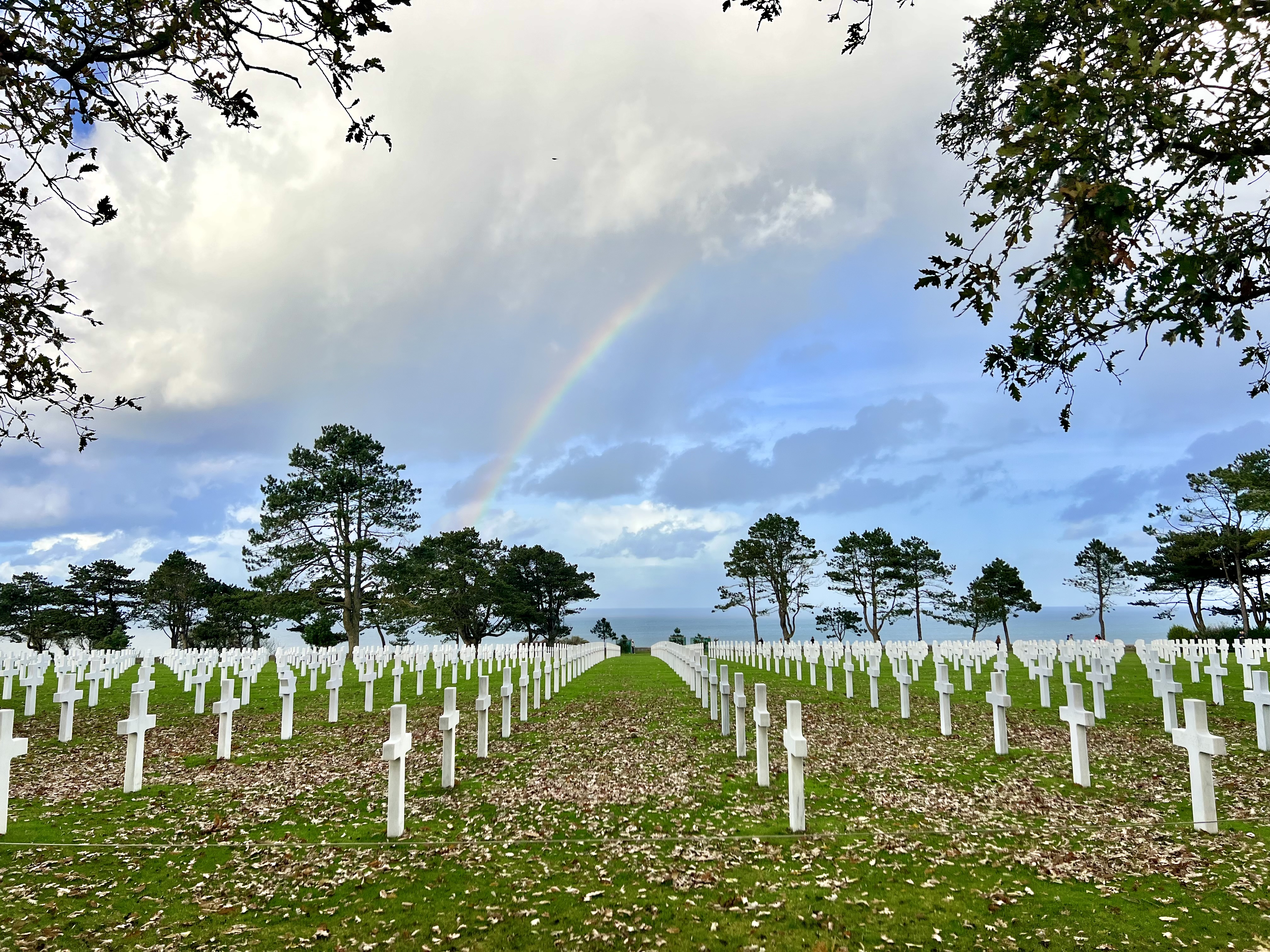 Rows of white crosses in a military cemetery stretch across a green lawn, with trees, a cloudy sky, and a faint rainbow overhead.